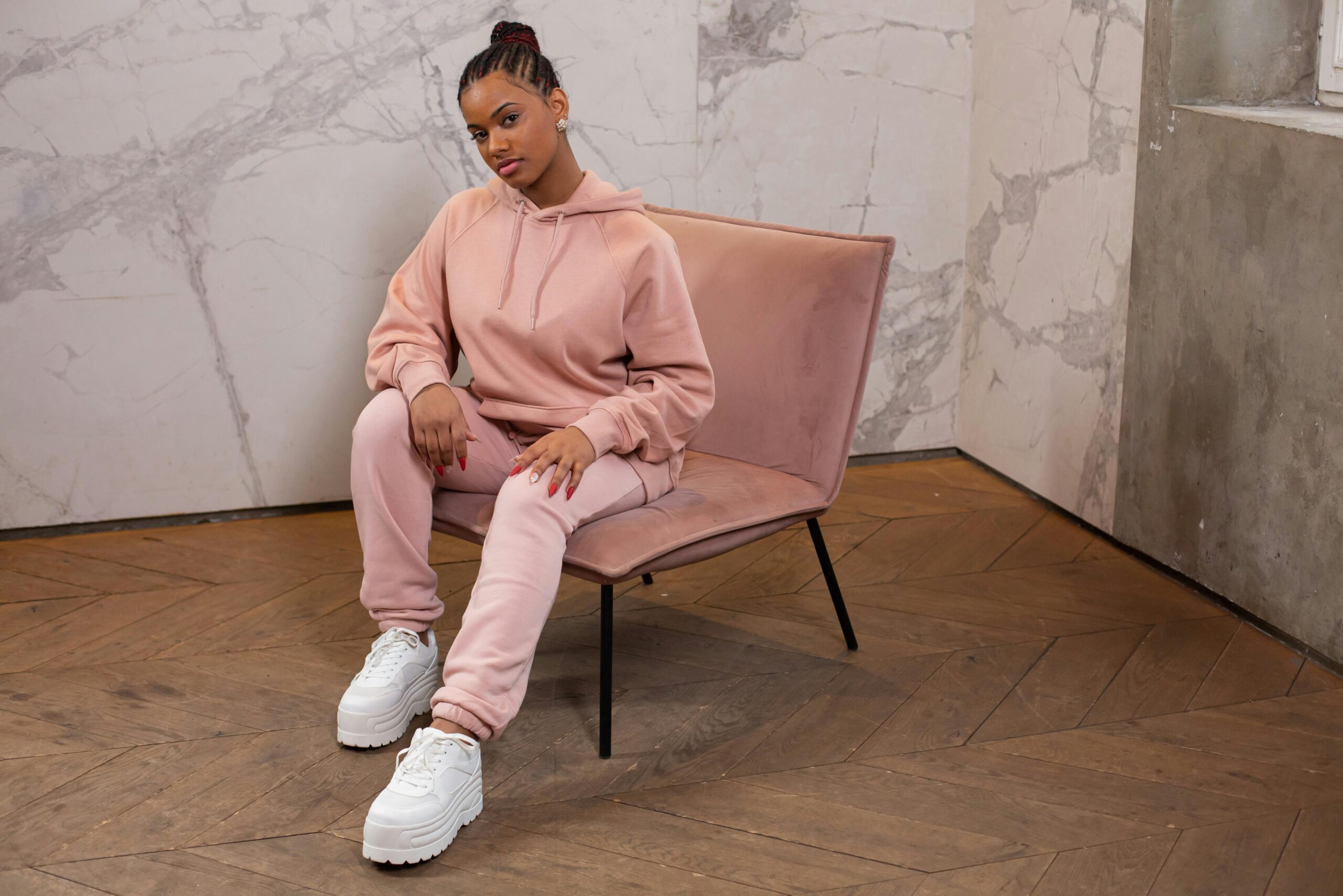 Full body of trendy African American female wearing stylish hoodie and pants looking at camera while sitting on armchair against wall